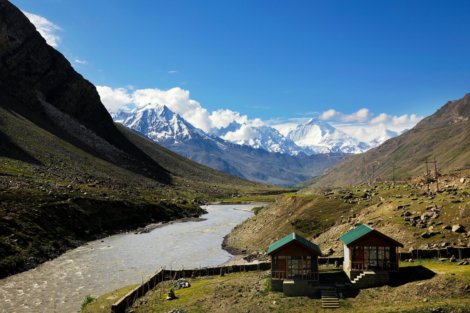 A serene mountain valley with a flowing river and cottages set against snow-covered Himalayan peaks in India.